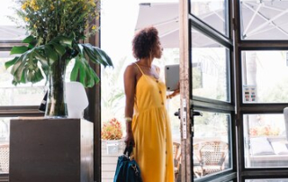 A stylish woman in a bright yellow dress holding a blue handbag stands at the open glass partition door of a modern entrance. The scene includes a black cabinet with a vase of fresh flowers and large outdoor umbrellas visible through the glass, creating an inviting and contemporary atmosphere