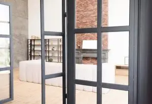 Contemporary office space featuring glass partition walls with black frames. The room includes a white sofa, exposed brick accent wall, and industrial-style shelving. The flooring is light wood, and the space is brightened by natural light filtering through large windows