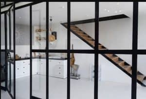 Contemporary kitchen area featuring sleek white cabinets, a floating wooden staircase, and glass partition walls with black frames. The space is accentuated with open shelving, gold accents, and minimalistic decor