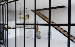 Contemporary kitchen area featuring sleek white cabinets, a floating wooden staircase, and glass partition walls with black frames. The space is accentuated with open shelving, gold accents, and minimalistic decor